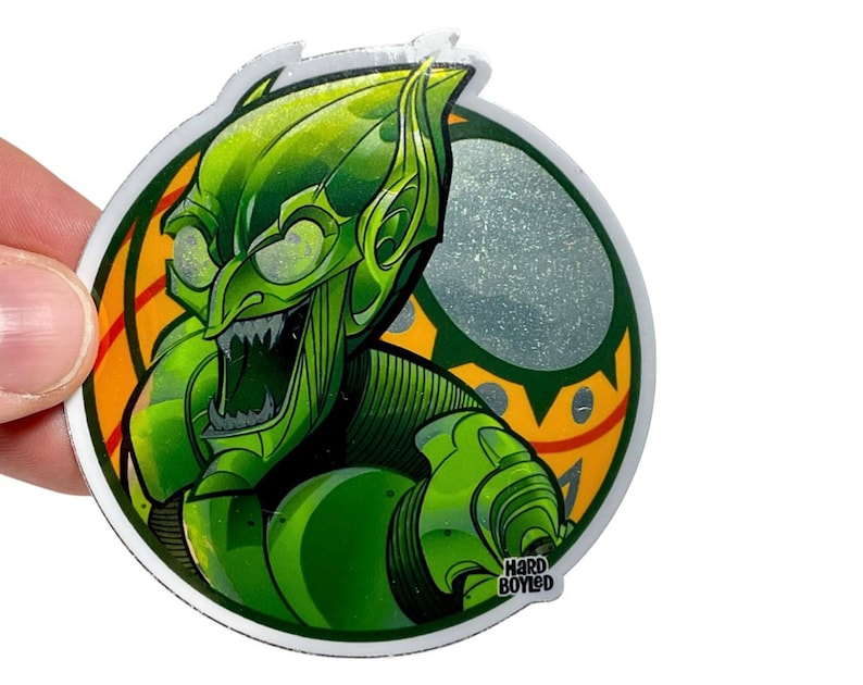 Puede incluir: Pegatina redonda verde y amarilla con una ilustraci&oacute;n de dibujos animados del Duende Verde de los c&oacute;mics de Spiderman. La pegatina tiene el texto "Hard Boiled" en la parte inferior.