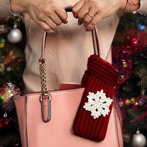 May include: A pink handbag with gold chain accents and a red knitted scarf with a white snowflake design. The scarf is hanging from the handbag. The background includes a Christmas tree with ornaments.