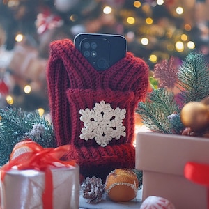 Puede incluir: Una funda roja de teléfono tejida con un diseño de copo de nieve blanco, que sostiene un teléfono inteligente negro. La funda se muestra con decoraciones navideñas, que incluyen una caja de regalo, adornos y ramas de pino, creando una escena festiva.