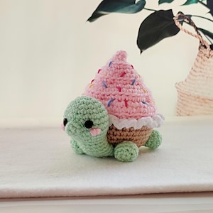 May include: A crocheted turtle with a pink cupcake shell. The turtle is light green with black eyes and pink cheeks. The cupcake shell is pink with colorful sprinkles and a white frilly edge. The turtle's shell is brown.