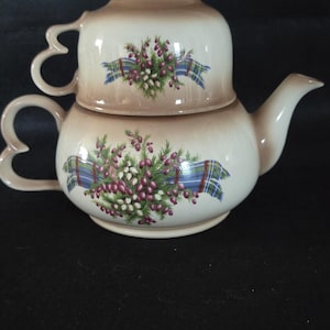 May include: A cream-coloured teapot and teacup set. The teapot has a curved spout and heart-shaped handle. Both pieces are decorated with a floral and berry design, accented with a blue and green tartan ribbon. A charming addition to any tea collection.