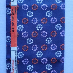 Puede incluir: Tela azul marino con un estampado floral repetido en blanco, naranja y morado. Una franja vertical blanca muestra "FR. FABRICS DESIGNER: MELODY MILLER" y "COLLECTION NAME AND DESCRIPTION: mustang".