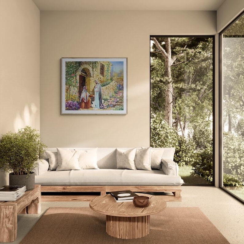 May include: A living room with a framed painting depicting figures near a stone building. A white sofa with pillows, a wooden coffee table, and a small side table with a plant are in the foreground. A large window offers a view of trees.