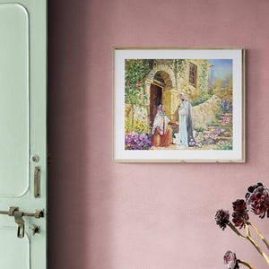 May include: A framed print depicting a religious scene with figures in flowing robes, set in a stone building with an arched doorway. The artwork features vibrant colors and is surrounded by a gold frame, mounted on a pink wall.