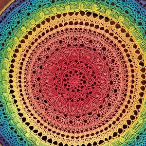 May include: A vibrant, circular crochet piece with a rainbow gradient, transitioning from blue to red. The intricate design features concentric circles with openwork patterns and a delicate, lacy texture. The piece is likely a decorative item, such as a doily or wall hanging.