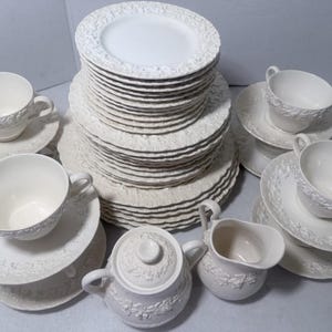 May include: A large set of off-white ceramic dinnerware featuring embossed floral and grape vine patterns along the rims. The collection includes a tall stack of dinner and salad plates, several teacups with matching saucers, a lidded sugar bowl, and a creamer.