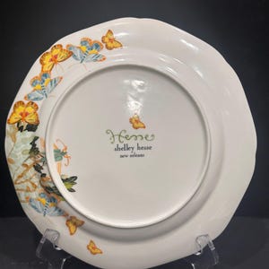 May include: White ceramic plate with a scalloped edge, decorated with yellow, blue, and orange butterflies. The plate has a floral design and the text "Hesse shelley hesse new orleans".