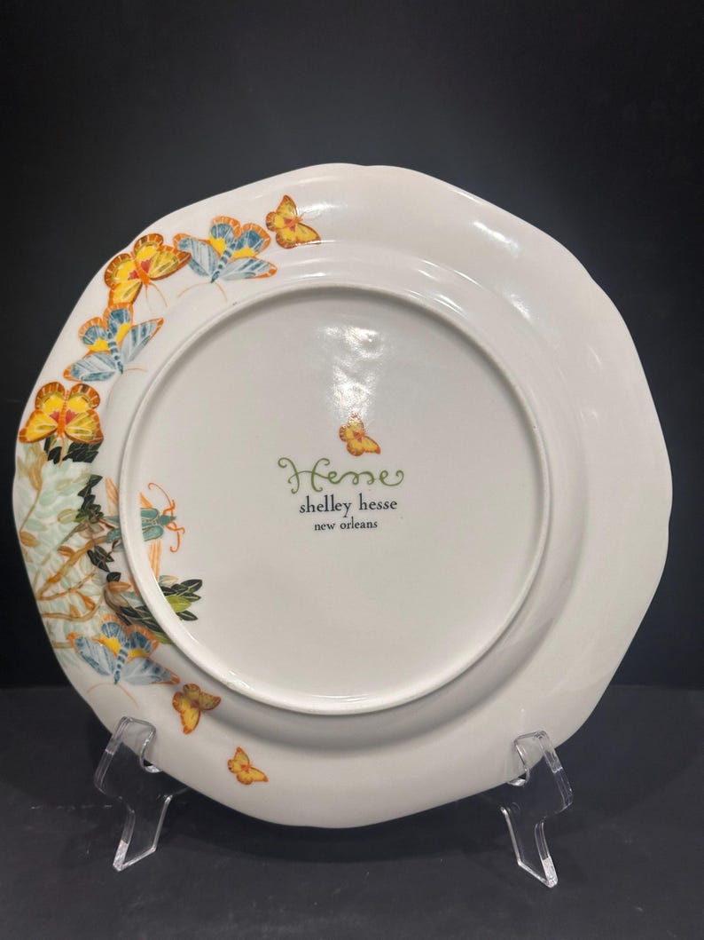 May include: White ceramic plate with a scalloped edge, decorated with yellow, blue, and orange butterflies. The plate has a floral design and the text "Hesse shelley hesse new orleans".