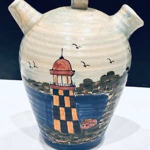 May include: Hand-painted ceramic pitcher with a lighthouse scene. The pitcher features a light blue and cream-colored body with a handle and two spouts. The lighthouse is painted with orange and black squares, with a red roof. Black birds are painted in the sky.