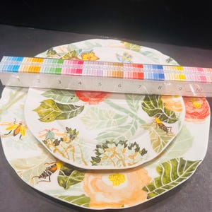 May include: Three white ceramic plates with floral and insect designs. The plates are stacked, with a ruler placed across them. The plates feature orange, yellow, and green floral patterns and insect illustrations.