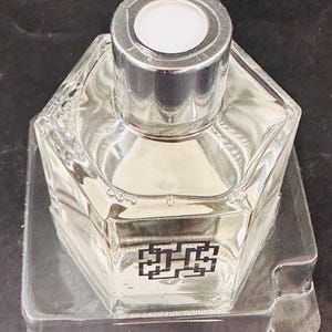 May include: A clear, hexagonal glass bottle with a silver cap and a white top. The bottle contains a light-colored liquid and sits in a clear plastic tray. A black and white graphic is on the front of the bottle.