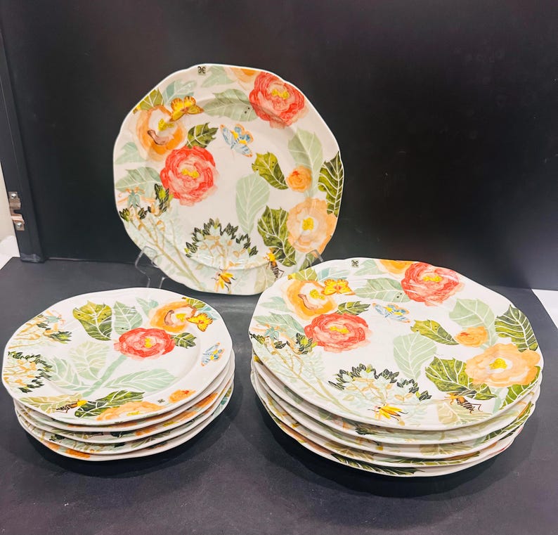 May include: A collection of ceramic plates with a floral design. The plates have a white base adorned with watercolour-style illustrations of red and orange flowers, green leaves, and small yellow details. The plates are in different sizes and shapes.