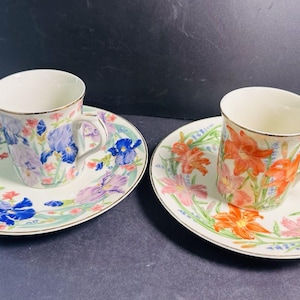 May include: Two ceramic tea cups and matching saucers. One set features blue and purple iris flower patterns, while the other set displays orange and pink lily flower designs. Both cups and saucers have a white base and thin gold-toned rims.