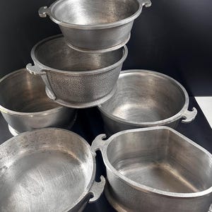 May include: A collection of six vintage metal cooking pots. The pots are silver-colored with textured exteriors and decorative handles. The pots vary in size and shape, with some having rounded edges and others more angular.