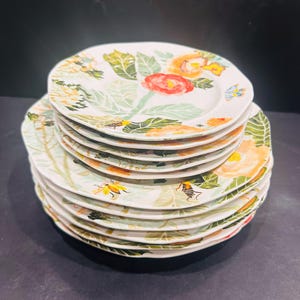 May include: A stack of white ceramic plates with a floral design. Each plate features a watercolour-style pattern of red, orange, and yellow flowers, green leaves, and small insects. The plates have a scalloped edge.