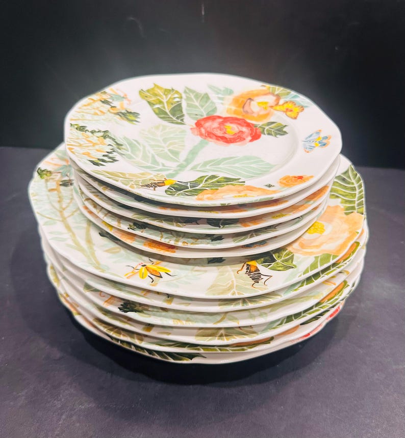 May include: A stack of white ceramic plates with a floral design. Each plate features a watercolour-style pattern of red, orange, and yellow flowers, green leaves, and small insects. The plates have a scalloped edge.