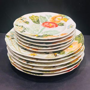 May include: A stack of white ceramic plates with a floral design. Each plate features a hand-painted pattern of red, orange, and yellow flowers, green leaves, and small insects. The plates have a slightly irregular, scalloped edge.