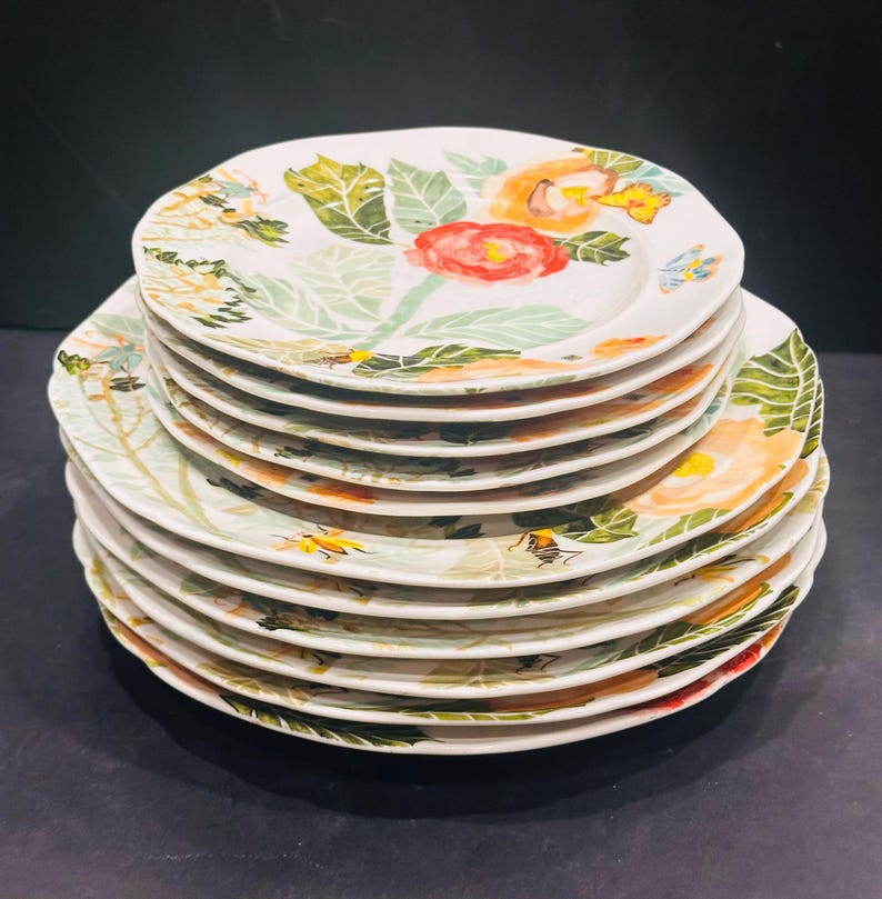 May include: A stack of white ceramic plates with a floral design. Each plate features a hand-painted pattern of red, orange, and yellow flowers, green leaves, and small insects. The plates have a slightly irregular, scalloped edge.