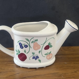 May include: A white ceramic watering can featuring a colorful fruit pattern including a red apple, a peach, a pear, blueberries, and blackberries with green vines. The watering can has a curved handle, a wide opening, and a spout with a blue rim.