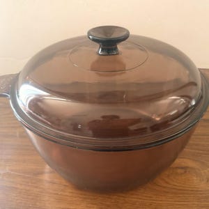 May include: A round, transparent amber-coloured glass casserole dish with two integrated side handles and a matching domed glass lid featuring a central knob handle. The cookware has a smooth, polished finish and a deep, wide bowl shape.