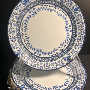 May include: A set of white ceramic plates with a blue floral design around the rim. The plates have a white center and a dark blue border. The floral pattern includes leaves and small flowers. The plates are stacked on top of each other.