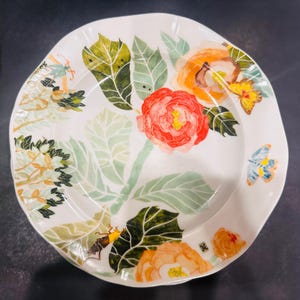 May include: White ceramic plate with a floral design featuring red, orange, and yellow flowers, green leaves, and butterflies. The plate has an irregular shape and is suitable for serving food.