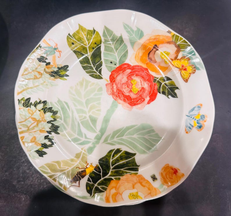 May include: White ceramic plate with a floral design featuring red, orange, and yellow flowers, green leaves, and butterflies. The plate has an irregular shape and is suitable for serving food.