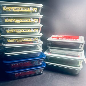 May include: A stack of rectangular glass food storage containers with light gray lids. Some containers feature a yellow Hello Kitty label. Other containers have blue lids and a Pyrex label. The containers are stacked on a dark surface.