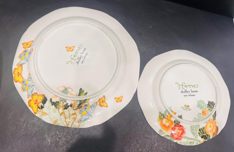 May include: Two white ceramic plates with scalloped edges. The plates are decorated with colourful floral and butterfly designs. The text "Heme shelley house new release" is printed on the plates.
