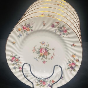 May include: A stack of white porcelain plates with a floral design and gold trim. Each plate features a central bouquet of pink, yellow, and purple flowers, with additional floral accents around the edges. The plates are displayed on a silver metal plate stand.
