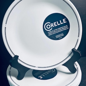 Puede incluir: Una pila de platos blancos Corelle con un borde azul marino y una pegatina con el logotipo azul marino. Los platos se exhiben en un soporte negro. La pegatina dice "VITRELLE EXTRA STRENGTH GLASS".
