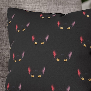 Könnte beinhalten: Schwarzes quadratisches Zierkissen mit einem sich wiederholenden Muster stilisierter schwarzer Katzenköpfe. Jedes Katzenkopf hat gelbe Augen und rosa Ohren. Das Kissen liegt auf einem grauen Polstersessel.