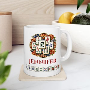 May include: A white ceramic mug featuring a colourful graphic of Mahjong tiles arranged in a circular pattern with floral accents. The name JENNIFER is printed in red capital letters below the graphic, followed by a row of individual Mahjong tiles.