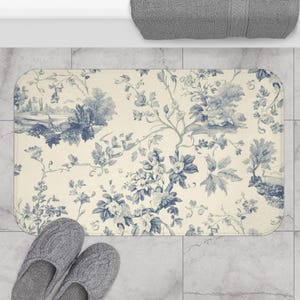 Könnte beinhalten: Eine rechteckige Badematte mit cremefarbenem Hintergrund und blauem Blumenmuster. Die Matte hat ein detailliertes Design aus Blumen, Bäumen und Landschaften. Graue Hausschuhe befinden sich am unteren Rand der Matte.