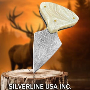 Puede incluir: Un cuchillo decorativo con una hoja estampada y un mango color crema con detalles dorados. El cuchillo se exhibe sobre un tocón de madera con el texto "SILVERLINE USA INC." en la parte inferior. El fondo presenta una imagen borrosa de un ciervo.