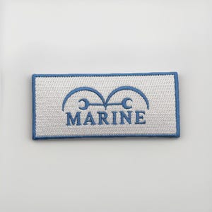 May include: Embroidered patch featuring the word "MARINE" in blue, with a stylized emblem above. The patch has a white background with a blue border. The design is rectangular.