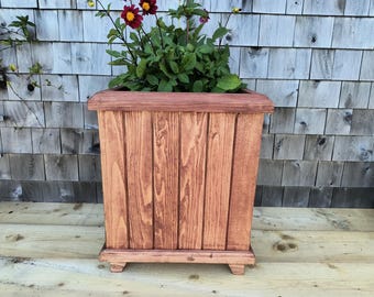 May include: A square, wooden planter box with a reddish-brown stain. The planter has a rounded top edge and is filled with green foliage and red flowers. The box is made of vertical wooden planks and sits on small feet.