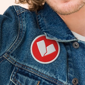 Puede incluir: Un parche redondo bordado con fondo rojo y diseño blanco. El parche está adherido a una chaqueta vaquera. El diseño presenta dos formas blancas, una más grande y otra más pequeña, con un borde curvo.