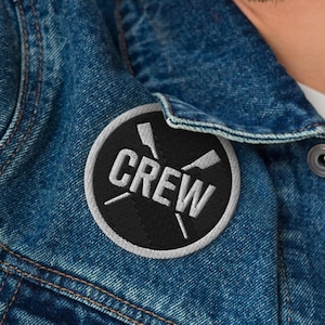 Könnte beinhalten: Runder, gestickter Aufnäher mit dem Wort "CREW" in Weiß. Er zeigt gekreuzte Ruder auf schwarzem Hintergrund mit grauem Rand. Der Aufnäher ist auf einer blauen Jeansjacke befestigt.