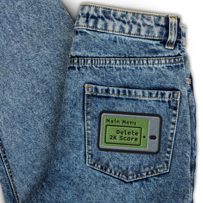 K&ouml;nnte beinhalten: Hellblaue Jeans mit einer Ges&auml;&szlig;tasche, die einen Aufn&auml;her hat. Der Aufn&auml;her zeigt einen gr&uuml;nen Bildschirm mit den wei&szlig;en Worten "Main Menu" und "Delete 2K Score", umrandet von einem schwarzen Rand.