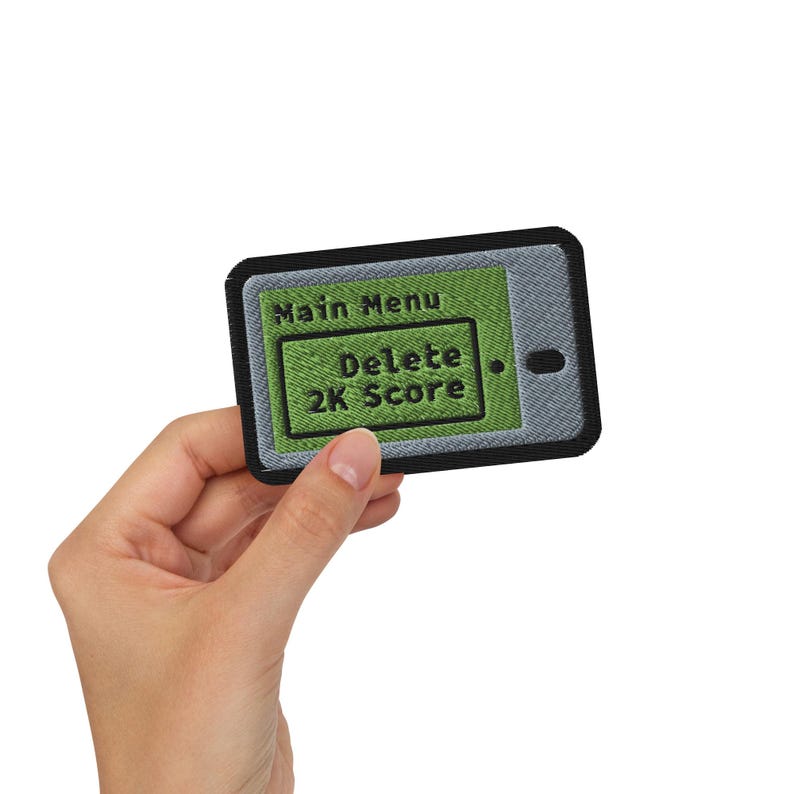 K&ouml;nnte beinhalten: Gestickter Aufn&auml;her in Form eines Smartphones, mit einem gr&uuml;nen Bildschirm mit dem Text "Main Menu" und "Delete 2K Score". Der Aufn&auml;her hat einen schwarzen Rand und einen grauen Hintergrund, der einem Telefonbildschirm &auml;hnelt. Auf der rechten Seite befindet sich ein kleiner, dunkler Punkt.