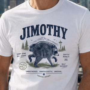 May include: A white short-sleeve t-shirt featuring a dark blue graphic of a raccoon with the text JIMOTHY. The design includes a mountain, the Space Needle, and text reading SHORT SPINE BIG LEGEND, SEATTLE ROYALTY, BUILT DIFFERENT, and UNBOTHERED. UNAPOLOGETIC. UNUSUAL. THRIVING ANYWAY.