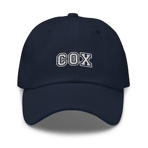 Könnte beinhalten: Marineblaue Baseballkappe mit dem Wort "COX" in weißen Blockbuchstaben gestickt. Die Kappe hat einen gebogenen Schirm und einen Stoffriemenverschluss auf der Rückseite. Ein klassisches, lässiges Accessoire.