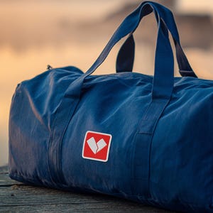 Puede incluir: Una bolsa de lona azul marino con asas resistentes y un parche cuadrado rojo con un gráfico blanco. La bolsa está hecha de una tela duradera y está diseñada para transportar artículos de viaje. La bolsa está sobre una superficie de madera.