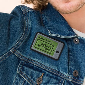 Puede incluir: Chaqueta vaquera azul con un parche rectangular. El parche tiene un diseño de pantalla verde con las palabras "Main Menu", "Delete" y "2K Score" en texto blanco. El parche es gris con un borde negro.