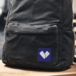 Puede incluir: Una mochila gris oscuro con un bolsillo frontal y un pequeño parche cuadrado. El parche es azul con un diseño blanco abstracto. La mochila está hecha de un material texturizado y tiene un cierre de cremallera en el bolsillo delantero.