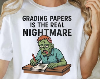 May include: White t-shirt with the text "GRADING PAPERS IS THE REAL NIGHTMARE." The shirt features a cartoon zombie grading papers at a desk. The zombie is green with exposed brain matter and wearing a blue shirt.