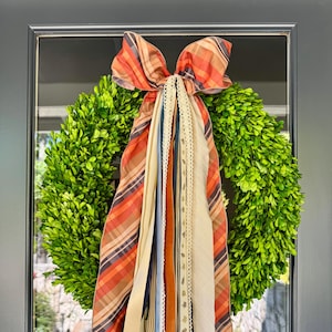 May include: A vibrant green boxwood wreath adorned with a large bow made of orange, brown, and cream striped fabric. Long, colorful ribbons in various textures and shades cascade down, adding a decorative touch to the wreath. A welcoming home decor item.