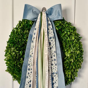 May include: A round, green boxwood wreath with a large, light blue velvet bow at the top. Several ribbons in various patterns and colors, including floral and striped designs, cascade down from the bow, adding a decorative touch.