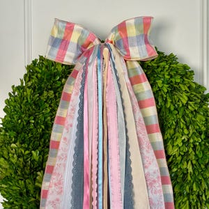 May include: A green boxwood wreath adorned with a large, pastel-colored plaid bow. The bow features long, flowing ribbons in various shades of pink, blue, and cream, with floral and lace patterns. A decorative accent for doors or walls.